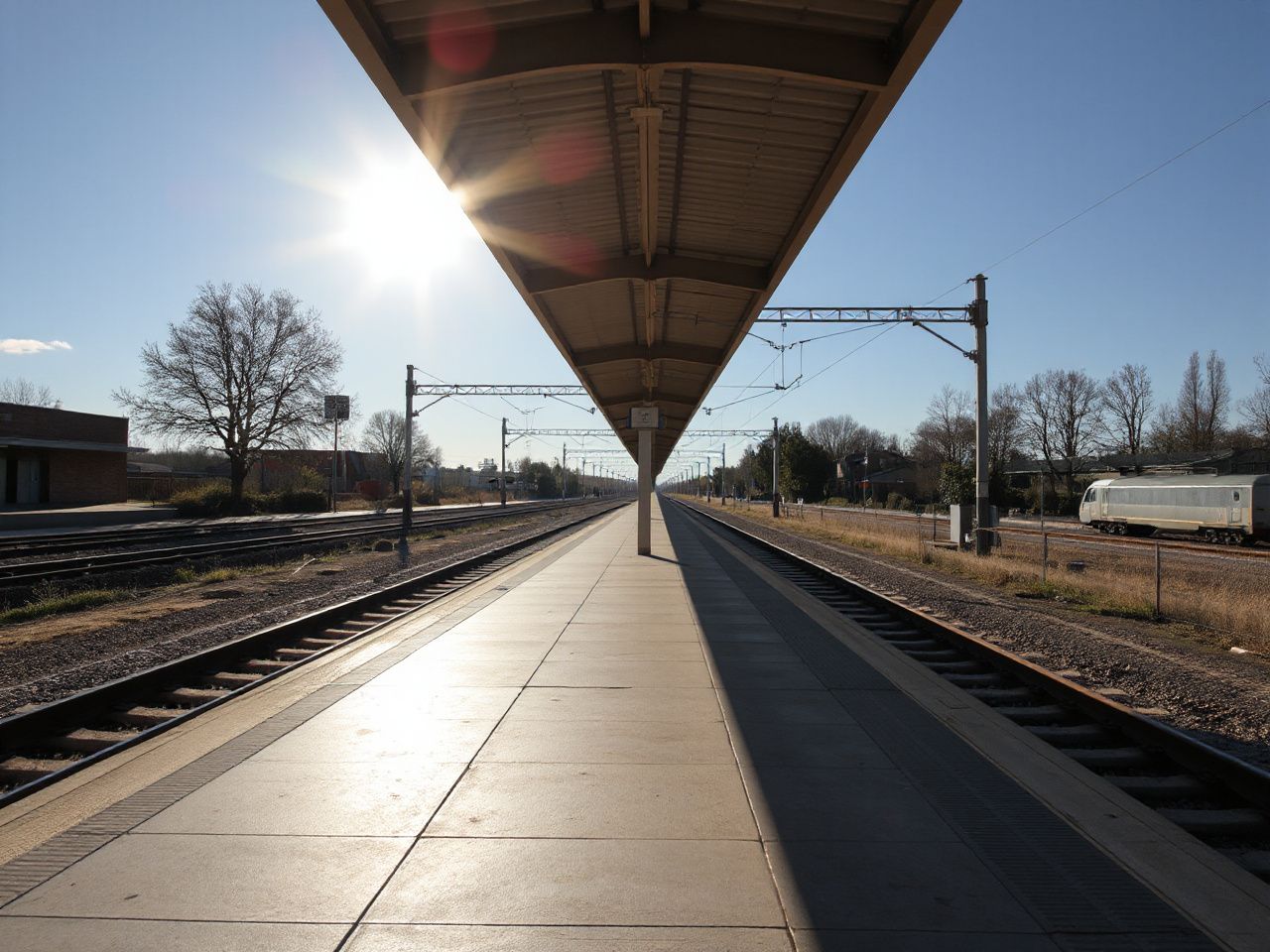 A sunlit train station platform with tracks stretching into the distance under a clear blue sky