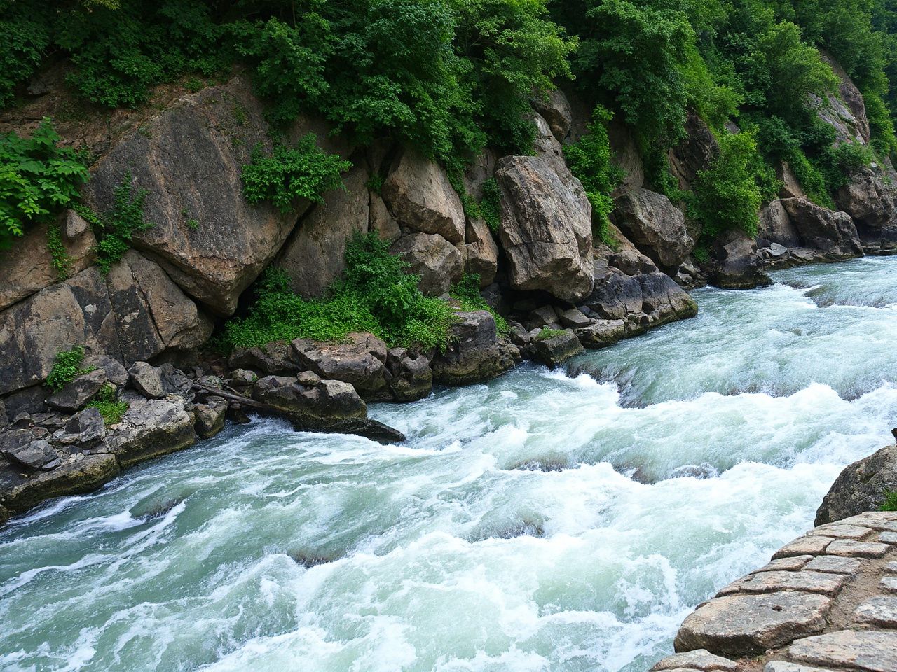 A rugged cliffside with rocky outcrops and lush greenery overlooking a fast-flowing river below