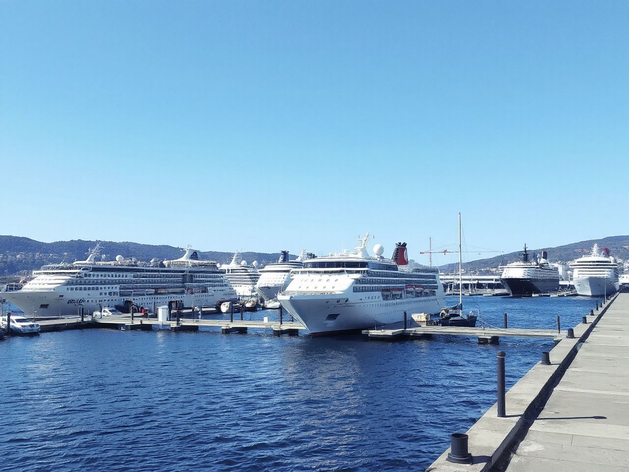 A bustling harbour with several cruise ships docked under clear blue skies, viewed from a nearby pier