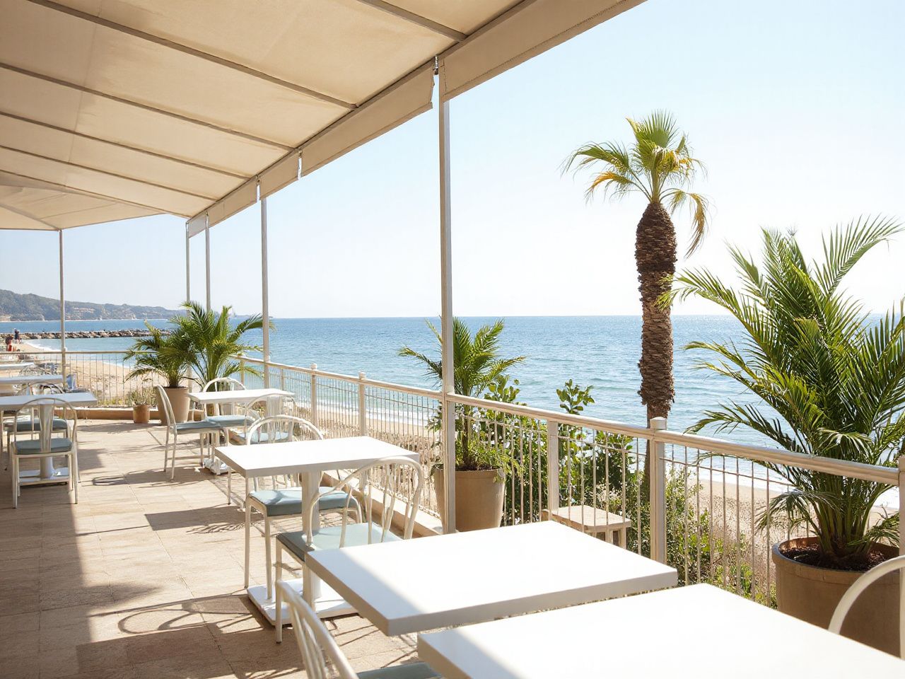 A sunlit beachside café terrace overlooking calm waters, with empty tables and tropical plants in the background