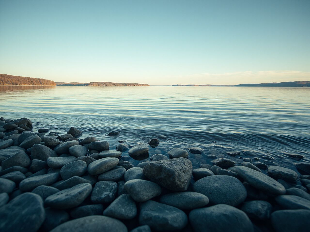 A serene lakeshore with smooth stones and gentle ripples reflecting the crisp fall sky at midday