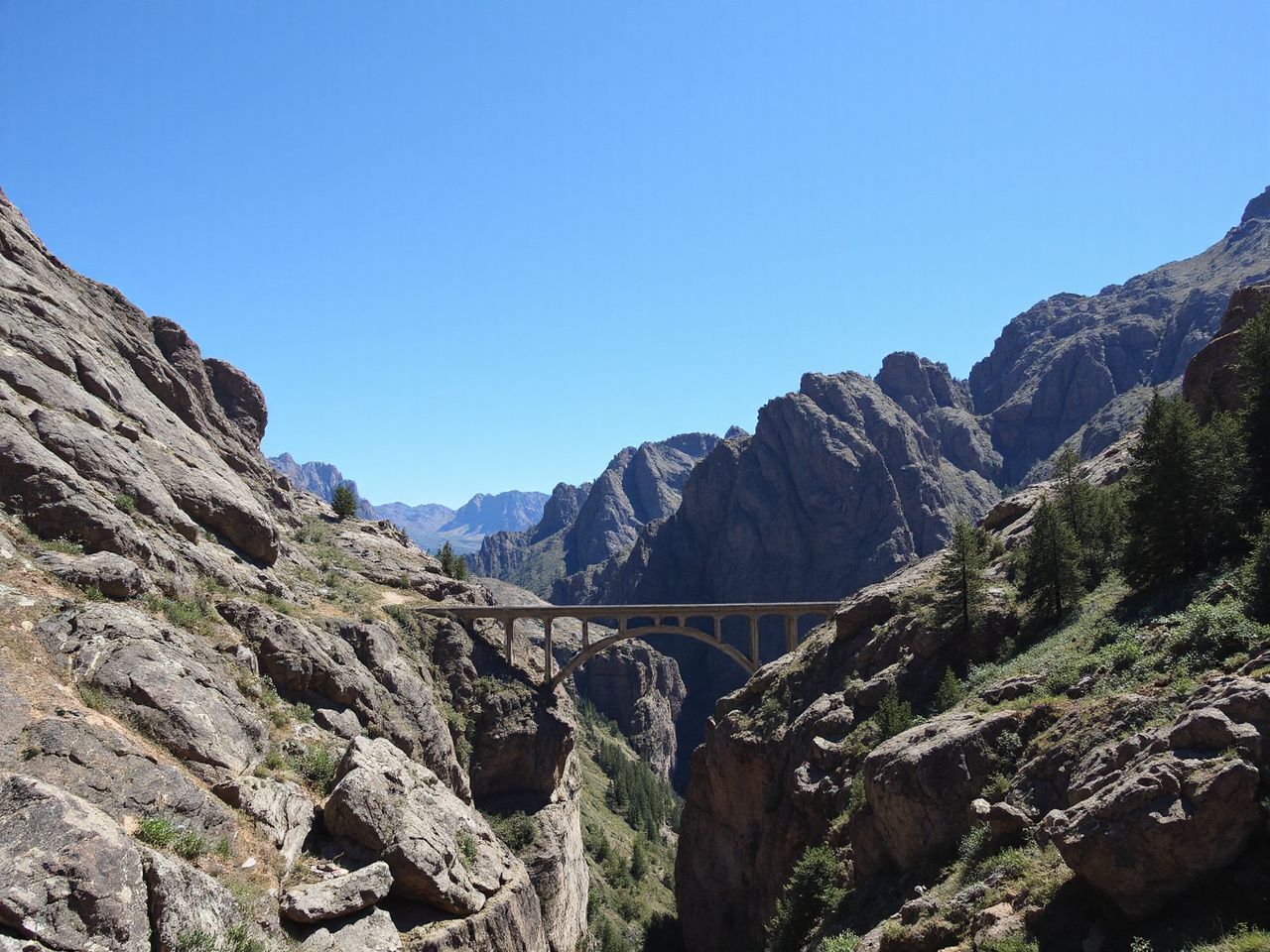 A rugged mountain landscape with a deep canyon and a narrow bridge spanning the gap under clear blue skies