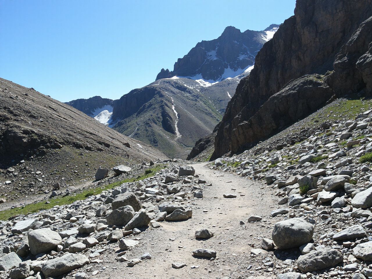 A rugged mountain trail winding through rocky terrain with scattered patches of snow and bright sunlight