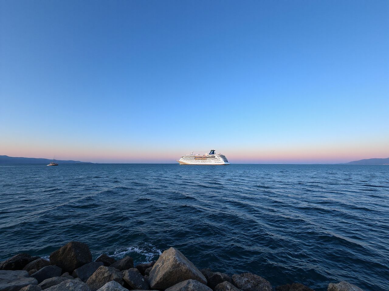 A large white cruise ship far out on a calm blue sea at sunset, seen small from a distant rocky shore