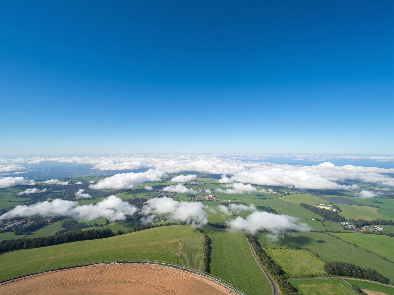 A wide blue sky over green farmland seen from very high up, with white clouds drifting far below