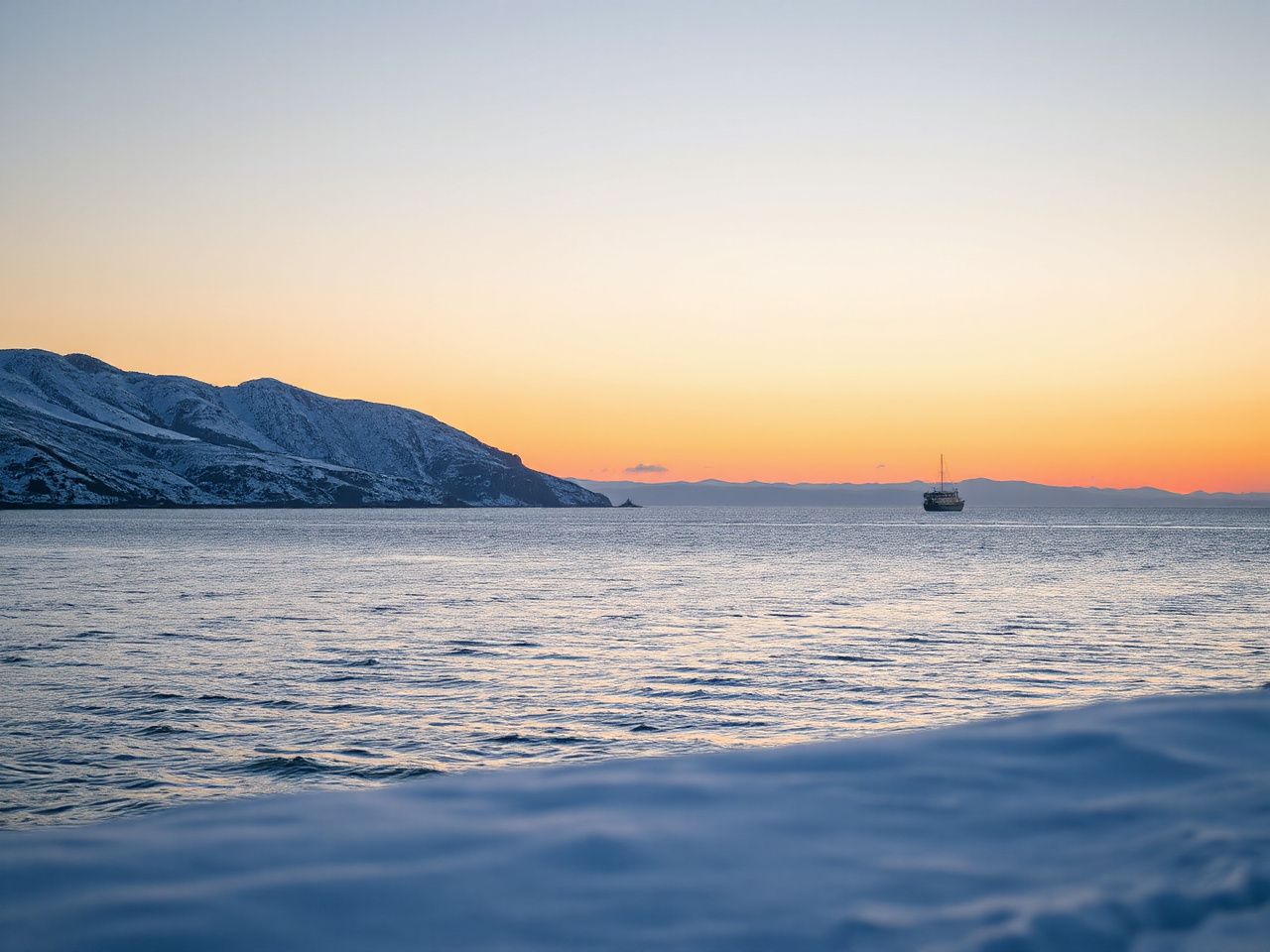 A calm sea at sunset beside snow-dusted coastal hills, with one small white ship far away on the horizon