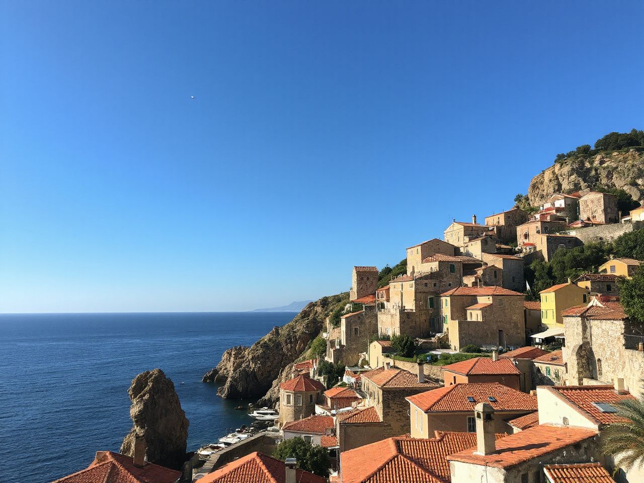 A sunlit coastal village with terracotta roofs nestled between rocky cliffs and a calm blue sea under a clear sky