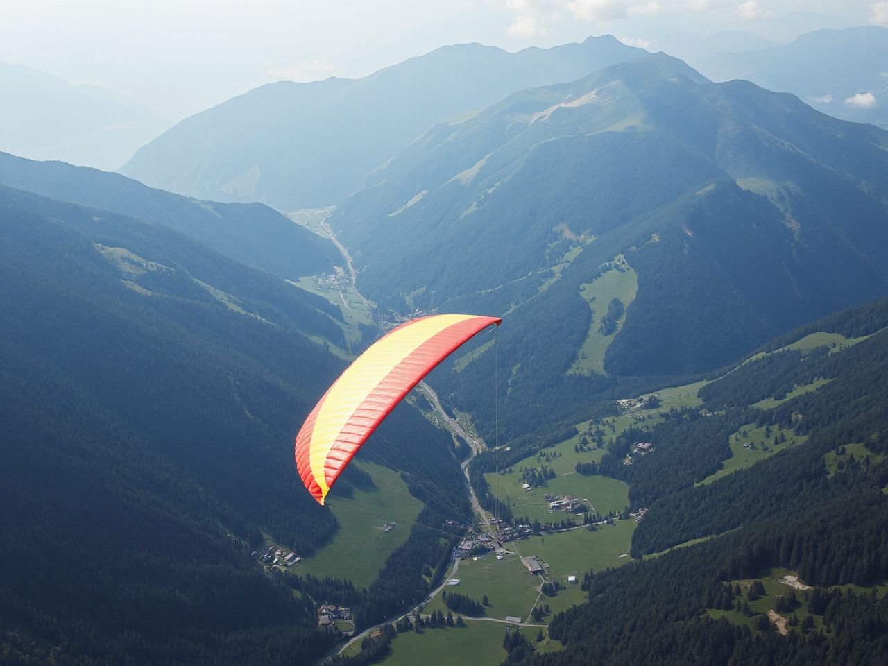 A red and yellow paraglider wing flying high above a green mountain valley, seen small from far away