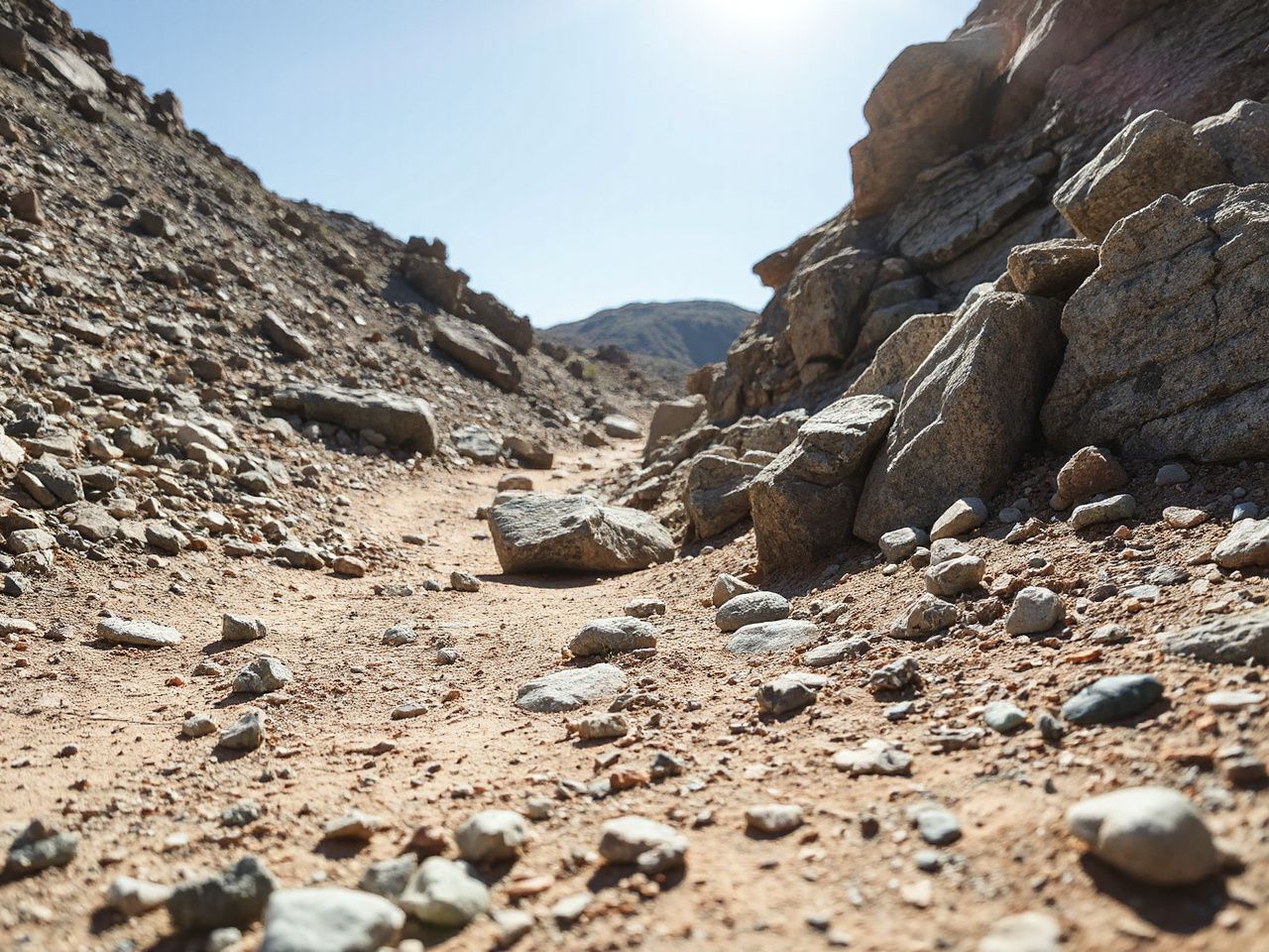 A close-up of a rugged mountain trail with loose rocks and patches of dirt under bright sunlight