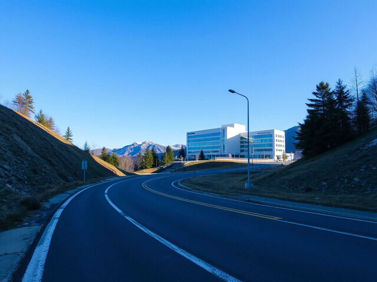 A quiet mountain road winding past a modern hospital building under a clear blue sky