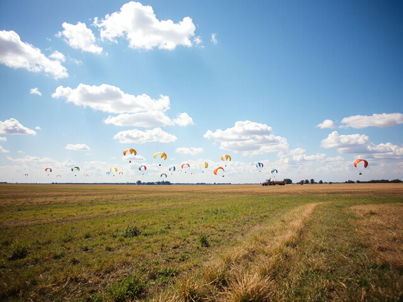 A bright blue sky with scattered clouds above an open field dotted with colorful parachutes gently descending