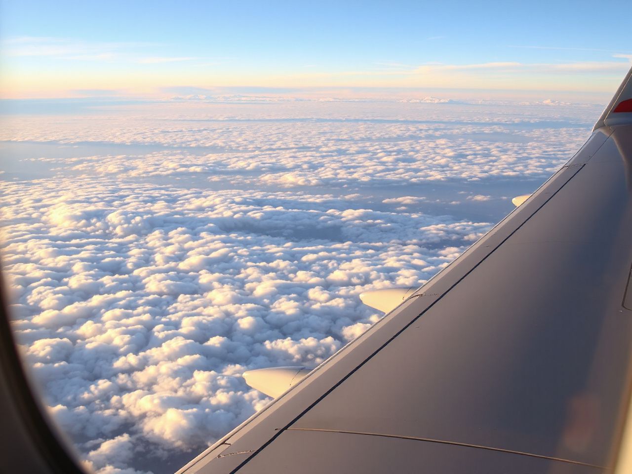 The wing of a passenger airplane above a sea of white clouds at sunrise, seen from a cabin window