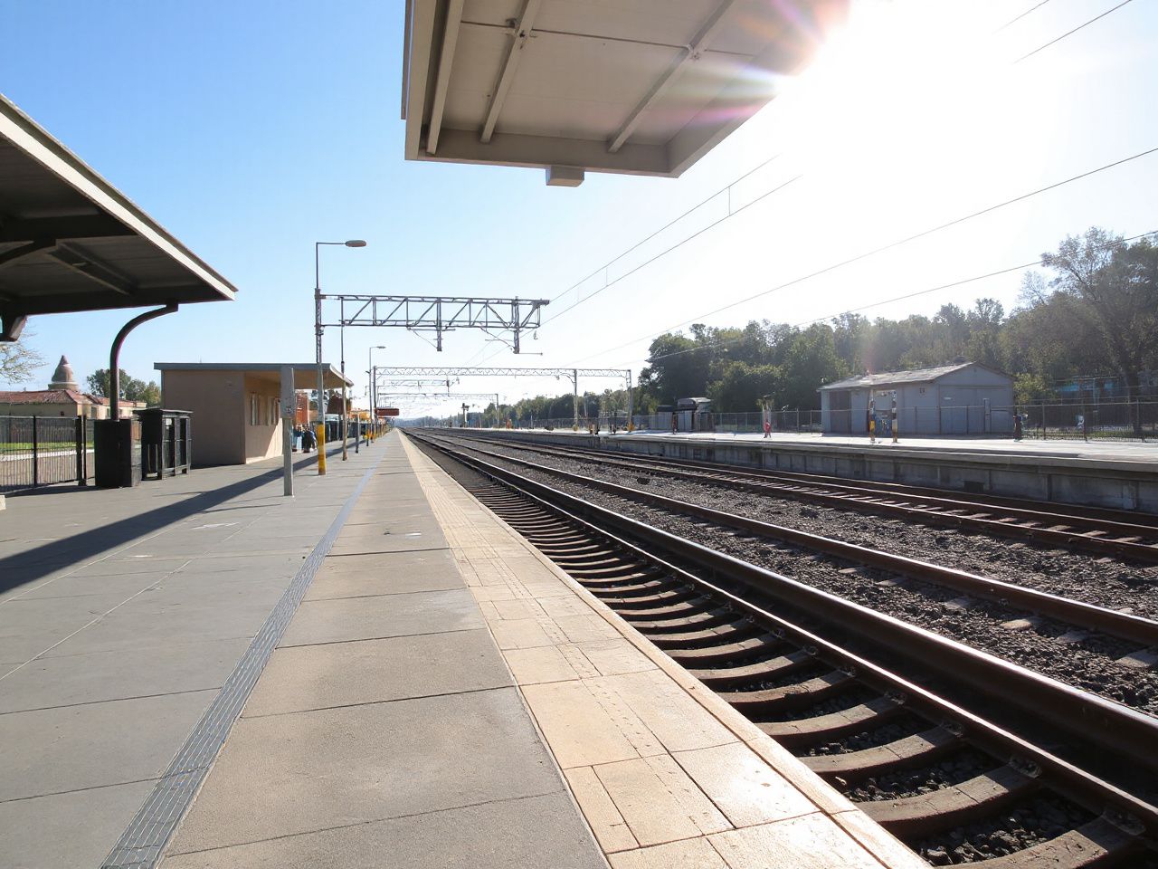 Sunlit train station platform with tracks stretching into the distance under a clear blue sky