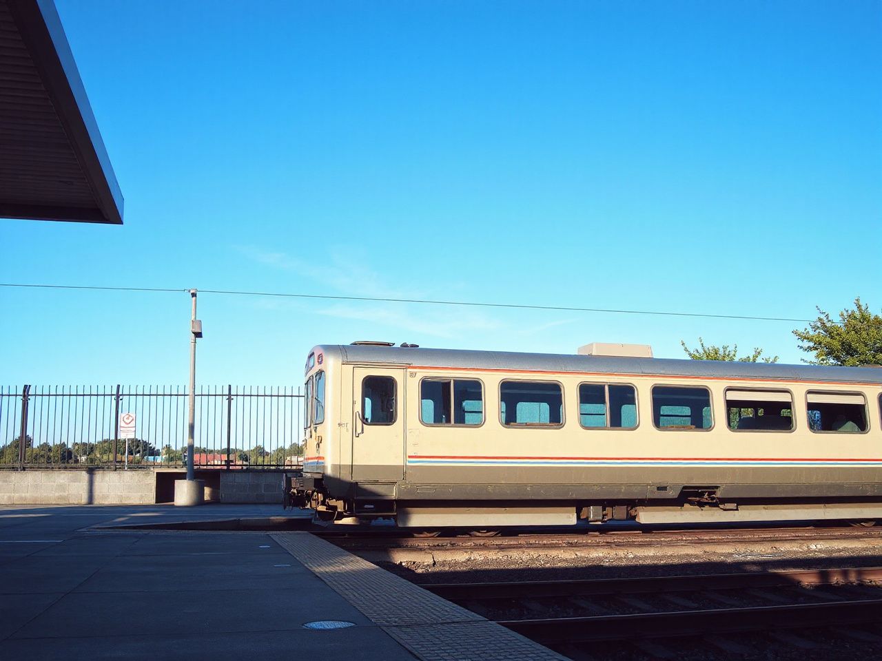 A sunlit station platform with a classic single-level passenger train waiting under clear blue skies