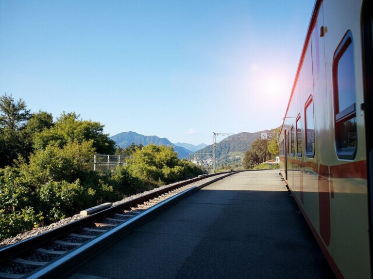 A sunlit train platform with distant mountains and lush greenery under a clear blue sky