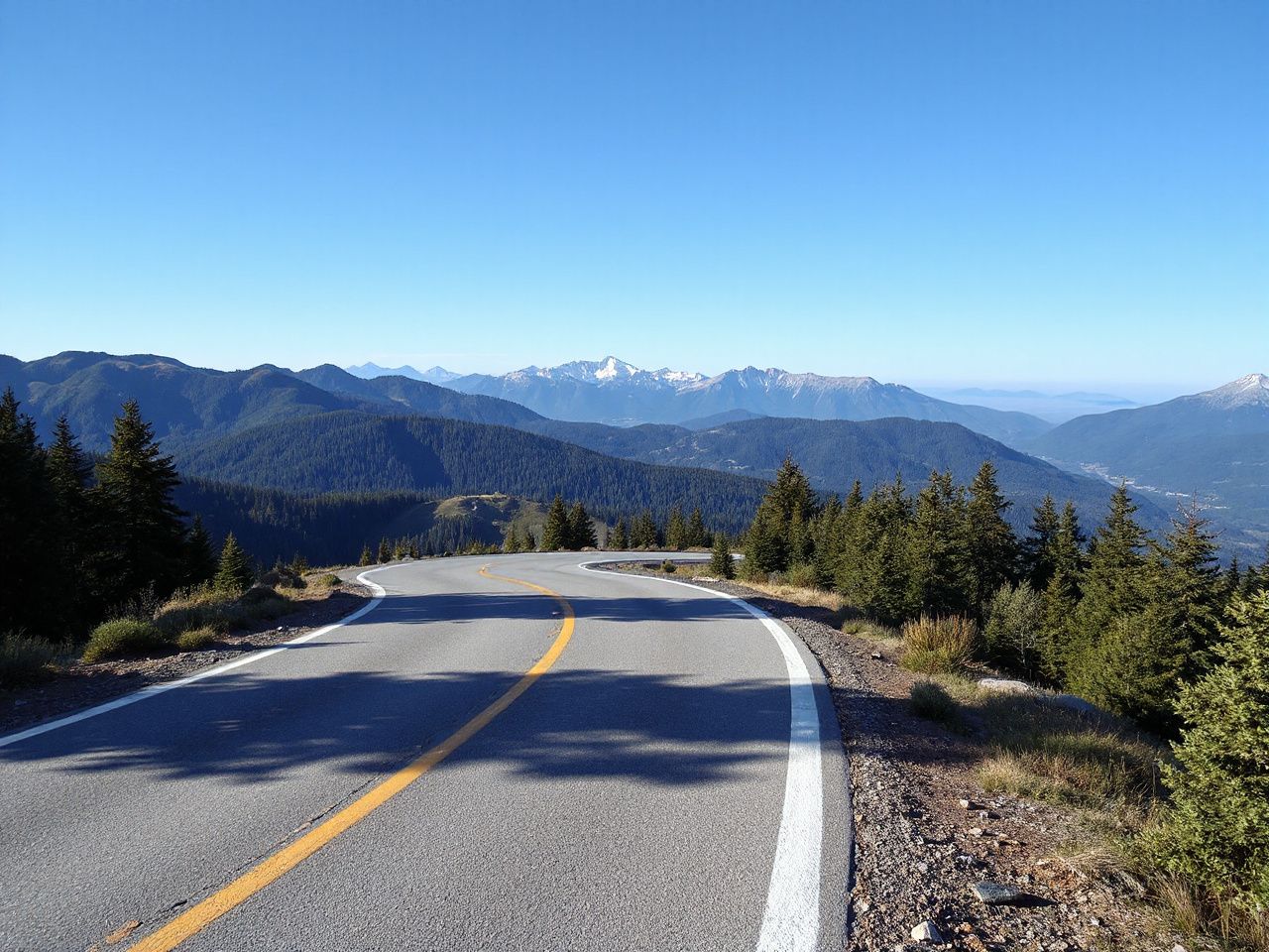 A winding mountain road under clear blue skies, surrounded by lush green forests and distant peaks